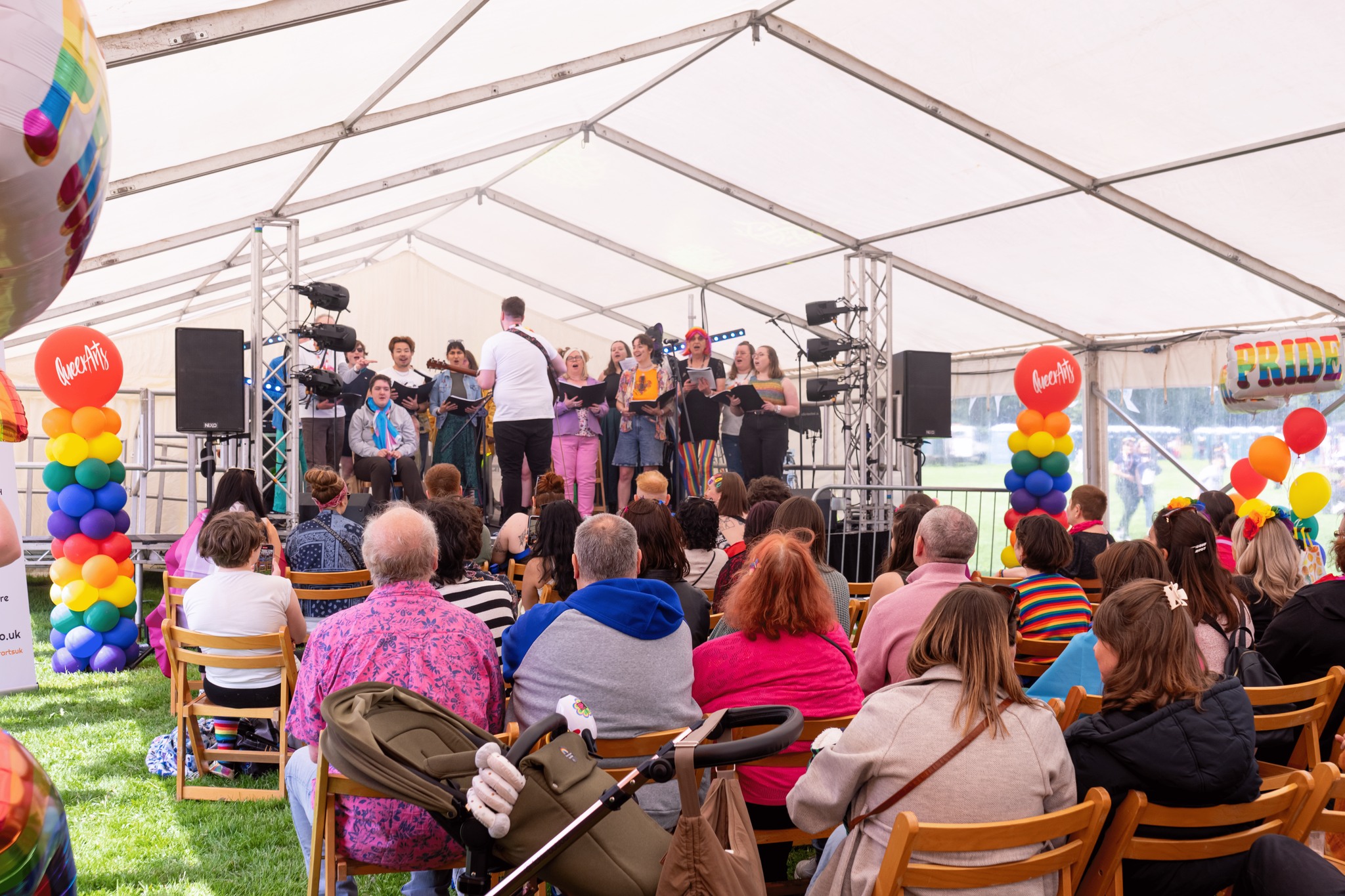 Choir performing on stage at York Pride, under a rainbow-decorated tent, with a diverse audience watching.