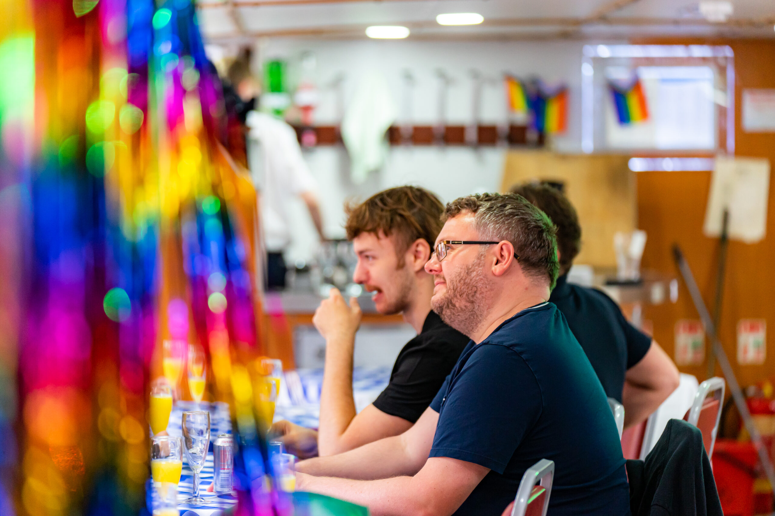 People enjoying drinks at a colorful party with vibrant rainbow decorations in the background.