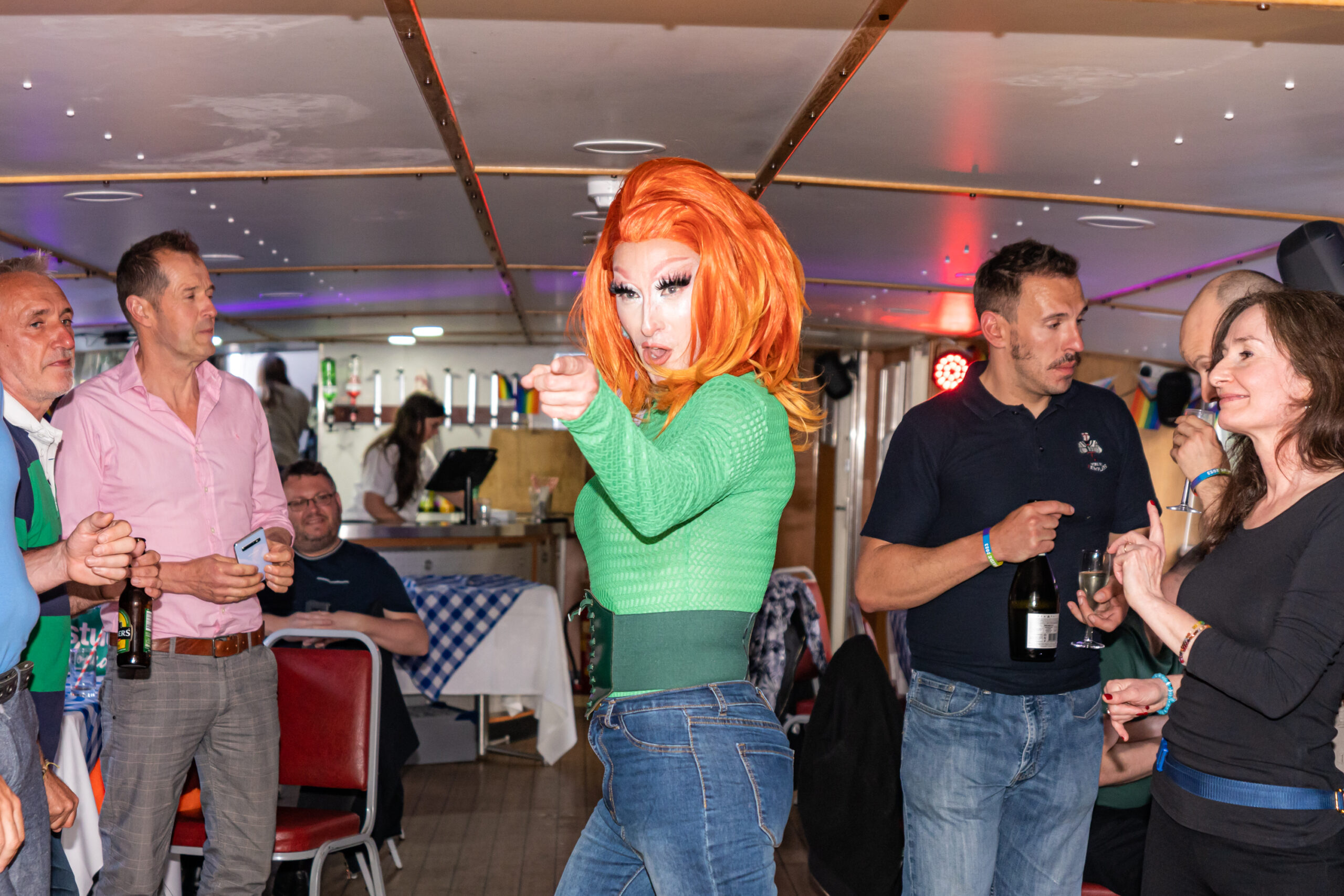Drag performer in vibrant wig entertains guests on a boat party, creating a lively atmosphere.