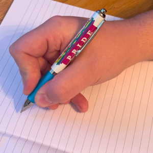 Hand holding a pen with Pride design, writing on a lined notebook.